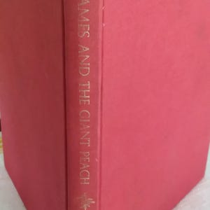 May include: A hardcover book with a solid red cover. The spine features gold lettering that reads James and the Giant Peach by Roald Dahl, accompanied by small gold leaf-like decorative symbols positioned above and below the title text.