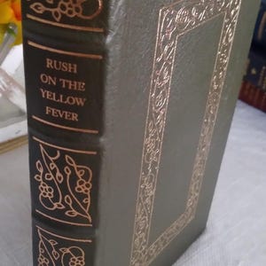 May include: A vintage book titled "Rush on the Yellow Fever" from The Classics of Medicine Library. The cover is dark green with gold embossed floral designs and text. The book's spine features the title and author in gold lettering.