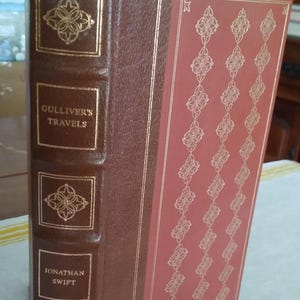 May include: A hardcover edition of "Gulliver's Travels" by Jonathan Swift. The book features a brown leather spine with gold embossed floral designs and text. The cover is a salmon color with a repeating gold pattern.