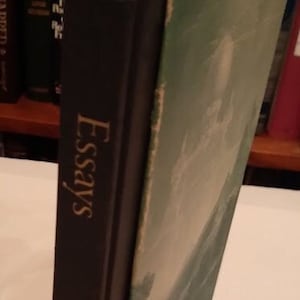 May include: A black hardcover book with the title "Essays" and the author "Emerson" printed in gold lettering. The book is in a slipcase with a green and white design.