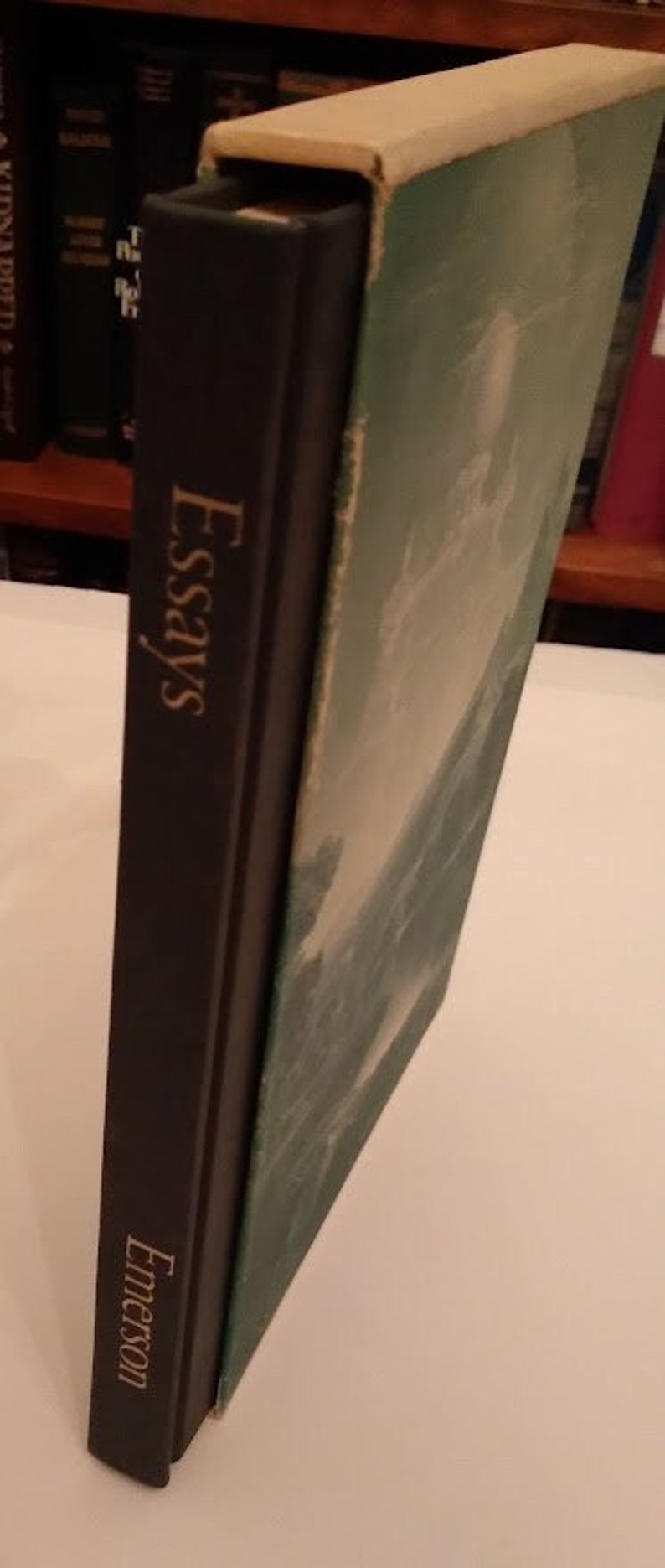 May include: A black hardcover book with the title "Essays" and the author "Emerson" printed in gold lettering. The book is in a slipcase with a green and white design.