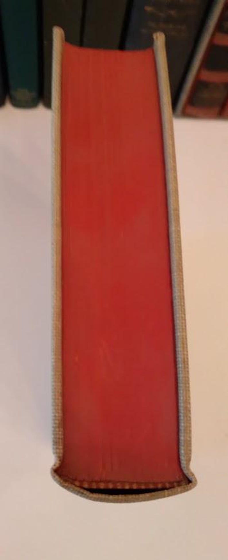 May include: A close-up of a book spine with a vibrant red cover. The edges are a light beige color, and the book is part of a collection. The spine is smooth and appears to be in good condition.