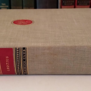 May include: A hardcover book titled "The Law of War and Peace" by Grotius. The book has a light tan linen cover with a red and gold spine. A red circular emblem is on the front cover. The Classics Club is printed on the spine.