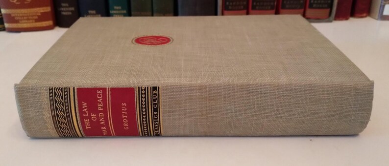 May include: A hardcover book titled "The Law of War and Peace" by Grotius. The book has a light tan linen cover with a red and gold spine. A red circular emblem is on the front cover. The Classics Club is printed on the spine.