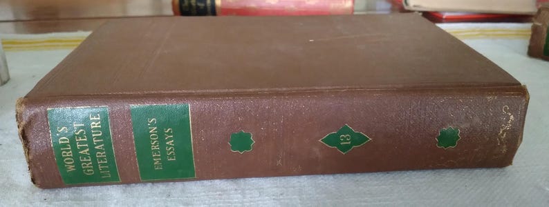 Pode incluir: Um livro de capa dura castanho com os t&iacute;tulos "World's Greatest Literature" e "Emerson's Essays" na lombada. A lombada apresenta etiquetas verdes com texto dourado e formas verdes decorativas. O n&uacute;mero "13" tamb&eacute;m &eacute; vis&iacute;vel.