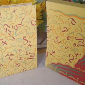 May include: Two hardcover books with yellow covers featuring a repeating pattern of stylized letters and symbols in red, orange, and teal. The book on the right includes a red and green graphic element at the bottom corner with wavy lines.
