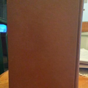 Pode incluir: Um livro de capa dura castanho est&aacute; em p&eacute; sobre uma superf&iacute;cie de madeira. A capa do livro &eacute; castanha lisa e mate, com uma lombada ligeiramente mais escura. As bordas das p&aacute;ginas s&atilde;o vis&iacute;veis e o livro parece estar em bom estado.