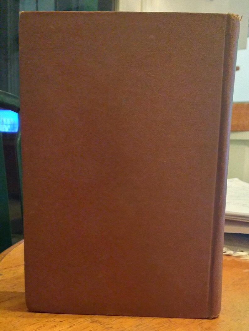Pode incluir: Um livro de capa dura castanho est&aacute; em p&eacute; sobre uma superf&iacute;cie de madeira. A capa do livro &eacute; castanha lisa e mate, com uma lombada ligeiramente mais escura. As bordas das p&aacute;ginas s&atilde;o vis&iacute;veis e o livro parece estar em bom estado.