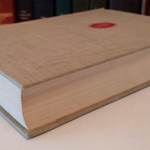 May include: A hardcover book with a beige linen cover and a red circular emblem. The book's pages are white, and the edges are beige. The book is thick, suggesting a substantial volume of text.