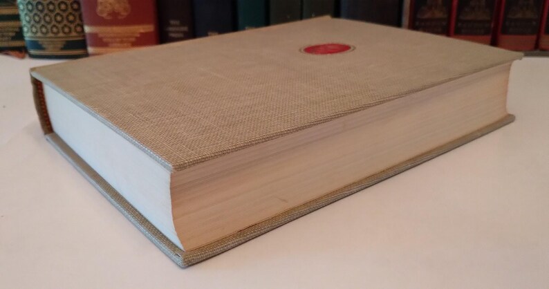 May include: A hardcover book with a beige linen cover and a red circular emblem. The book's pages are white, and the edges are beige. The book is thick, suggesting a substantial volume of text.