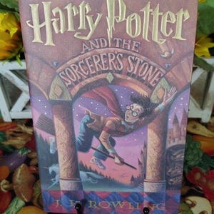 May include: A hardcover book titled Harry Potter and the Sorcerer's Stone by J.K. Rowling. The cover features a central illustration of a young person with glasses flying on a broomstick through a stone archway, with a unicorn visible below.