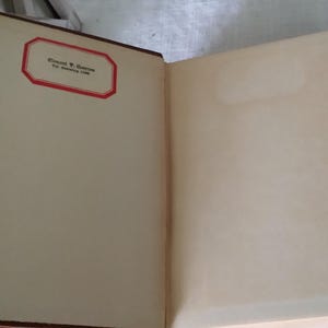 Pode incluir: Livro antigo aberto com p&aacute;ginas amareladas. A p&aacute;gina esquerda apresenta um r&oacute;tulo com borda vermelha e texto. A lombada &eacute; marrom escuro. O livro est&aacute; aberto, revelando as p&aacute;ginas internas.