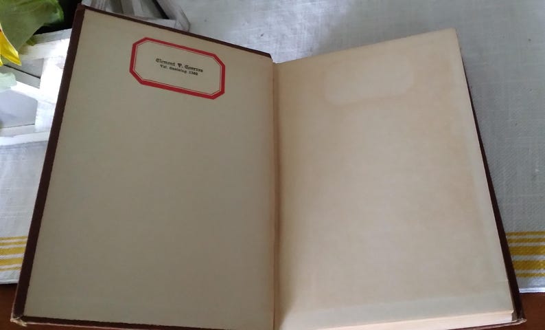 Pode incluir: Livro antigo aberto com p&aacute;ginas amareladas. A p&aacute;gina esquerda apresenta um r&oacute;tulo com borda vermelha e texto. A lombada &eacute; marrom escuro. O livro est&aacute; aberto, revelando as p&aacute;ginas internas.