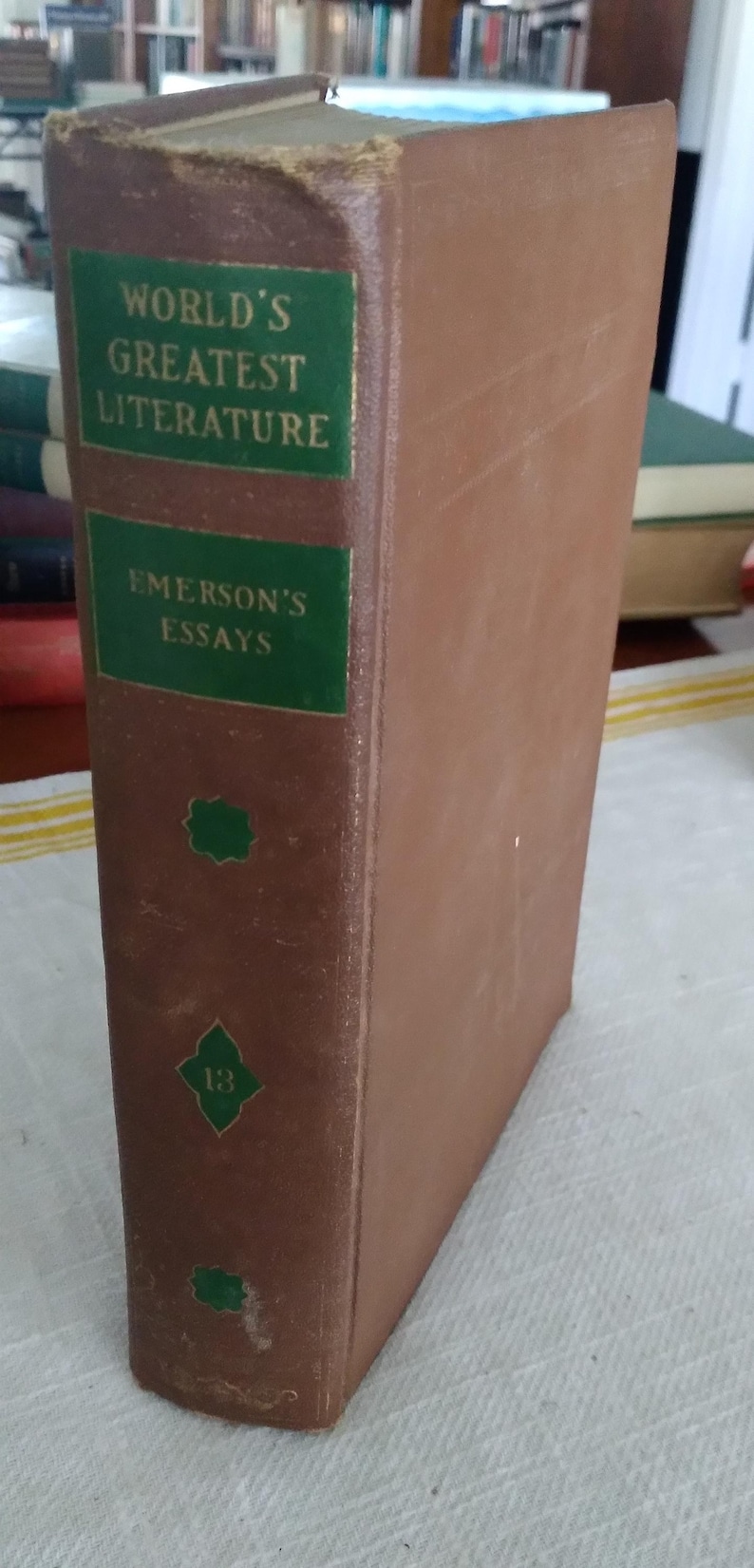 Pode incluir: Um livro antigo de capa dura intitulado "World's Greatest Literature" e "Emerson's Essays". O livro tem uma capa castanha com texto verde e elementos decorativos. A lombada desgastada indica a sua idade.