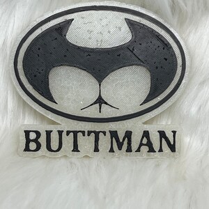 May include: A white 3D printed Batman logo with a black outline and the word "BUTTMAN" printed below it.
