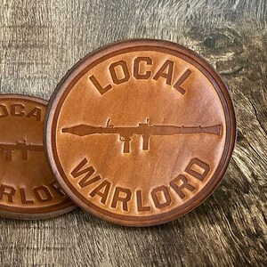 May include: Four brown leather coasters with the text "LOCAL WARLORD" and a silhouette of a rifle printed on them.