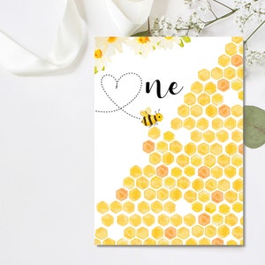 Puede incluir: Una tarjeta blanca con un patr&oacute;n de panal amarillo y una abeja volando hacia la palabra "uno" con un coraz&oacute;n encima.