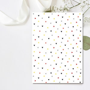 May include: White paper with a repeating pattern of small triangles in black, red, yellow, and gray. A white ribbon and sprigs of white flowers and a green leaf are in the background.