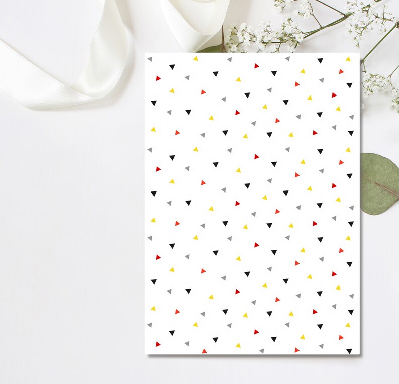 May include: White paper with a repeating pattern of small triangles in black, red, yellow, and gray. A white ribbon and sprigs of white flowers and a green leaf are in the background.