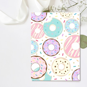 May include: A white card featuring a pattern of colorful donuts with sprinkles. The donuts have pink, blue, purple, and brown frosting. Some donuts have a bite taken out of them. Gold confetti dots are scattered around the donuts.