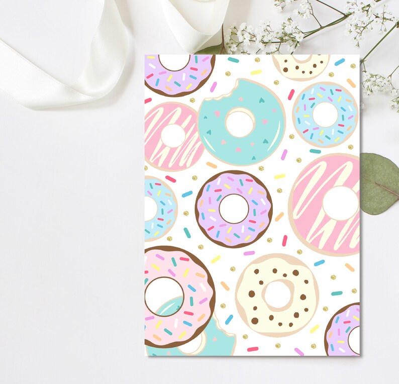 May include: A white card featuring a pattern of colorful donuts with sprinkles. The donuts have pink, blue, purple, and brown frosting. Some donuts have a bite taken out of them. Gold confetti dots are scattered around the donuts.