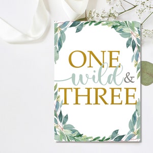 Peut inclure: Une affiche rectangulaire blanche avec l'inscription "ONE wild & THREE" en or et bleu clair. L'affiche est encadr&eacute;e d'une bordure de feuilles et de verdure aquarelles vertes et bleues.
