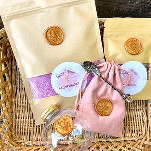 May include: A woven basket holds several items, including a pink drawstring bag, a clear glass container, and pouches sealed with wax stamps. The pouches contain a purple powder and other ingredients. The text "Fairy Magic" is visible.