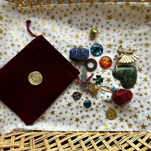 K&ouml;nnte beinhalten: Eine Sammlung dekorativer Verzierungen, darunter ein bordeauxfarbener Samtbeutel mit einem goldenen Siegel, auf sternf&ouml;rmigem Papier in einem geflochtenen Korb angeordnet. Die Verzierungen umfassen einen blauen Glitzerw&uuml;rfel, einen goldenen W&uuml;rfel und verschiedene Charms.