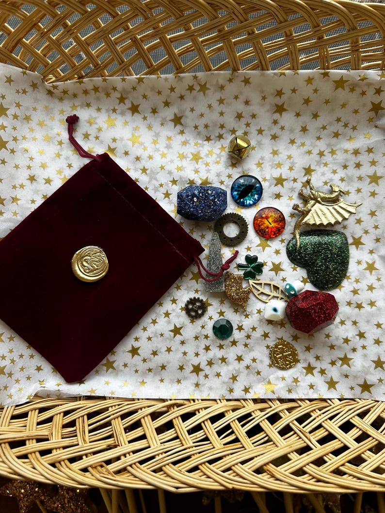K&ouml;nnte beinhalten: Eine Sammlung dekorativer Verzierungen, darunter ein bordeauxfarbener Samtbeutel mit einem goldenen Siegel, auf sternf&ouml;rmigem Papier in einem geflochtenen Korb angeordnet. Die Verzierungen umfassen einen blauen Glitzerw&uuml;rfel, einen goldenen W&uuml;rfel und verschiedene Charms.