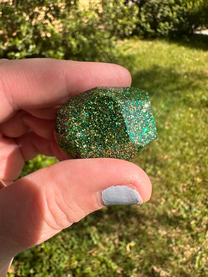 K&ouml;nnte beinhalten: Ein facettiertes, geometrisches Objekt, das mit gr&uuml;nem Glitzer bedeckt ist. Das Objekt wird in einer Hand vor dem Hintergrund von gr&uuml;nem Gras und Sonnenlicht gehalten. Der Glitzer f&auml;ngt das Licht ein und erzeugt einen funkelnden Effekt.