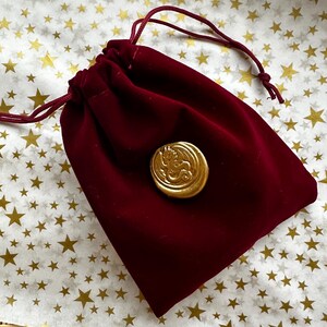 K&ouml;nnte beinhalten: Ein weinroter Samtbeutel mit Kordelzug und einem goldfarbenen Knopf mit einem Drachendesign. Der Beutel wird mit einer passenden roten Kordel verschlossen. Der Hintergrund ist eine wei&szlig;e Oberfl&auml;che mit goldenen Sternmustern.