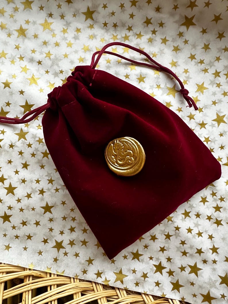 K&ouml;nnte beinhalten: Ein weinroter Samtbeutel mit Kordelzug und einem goldfarbenen Knopf mit einem Drachendesign. Der Beutel wird mit einer passenden roten Kordel verschlossen. Der Hintergrund ist eine wei&szlig;e Oberfl&auml;che mit goldenen Sternmustern.