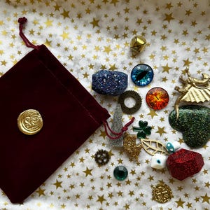 K&ouml;nnte beinhalten: Eine Sammlung dekorativer Verzierungen, darunter ein bordeauxfarbener Samtbeutel mit einem goldenen Siegel, glitzernde geometrische Formen, Zahnr&auml;der und augenf&ouml;rmige Cabochons. Die Artikel sind auf Papier mit Sternenmuster angeordnet.