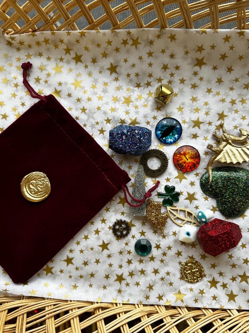 K&ouml;nnte beinhalten: Eine Sammlung dekorativer Verzierungen, darunter ein bordeauxfarbener Samtbeutel mit einem goldenen Siegel, glitzernde geometrische Formen, Zahnr&auml;der und augenf&ouml;rmige Cabochons. Die Artikel sind auf Papier mit Sternenmuster angeordnet.