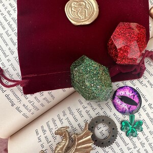 K&ouml;nnte beinhalten: Eine Sammlung von Fantasy-Accessoires, darunter ein goldener Drache, ein goldener Wachssiegel, ein roter Glitzer-Edelstein, ein gr&uuml;ner Glitzer-Edelstein, ein lila Augen-Cabochon, ein Zahnrad und ein gr&uuml;nes Kleeblatt. Diese Artikel sind auf einem offenen Buch und einem bordeauxroten Samtbeutel angeordnet.