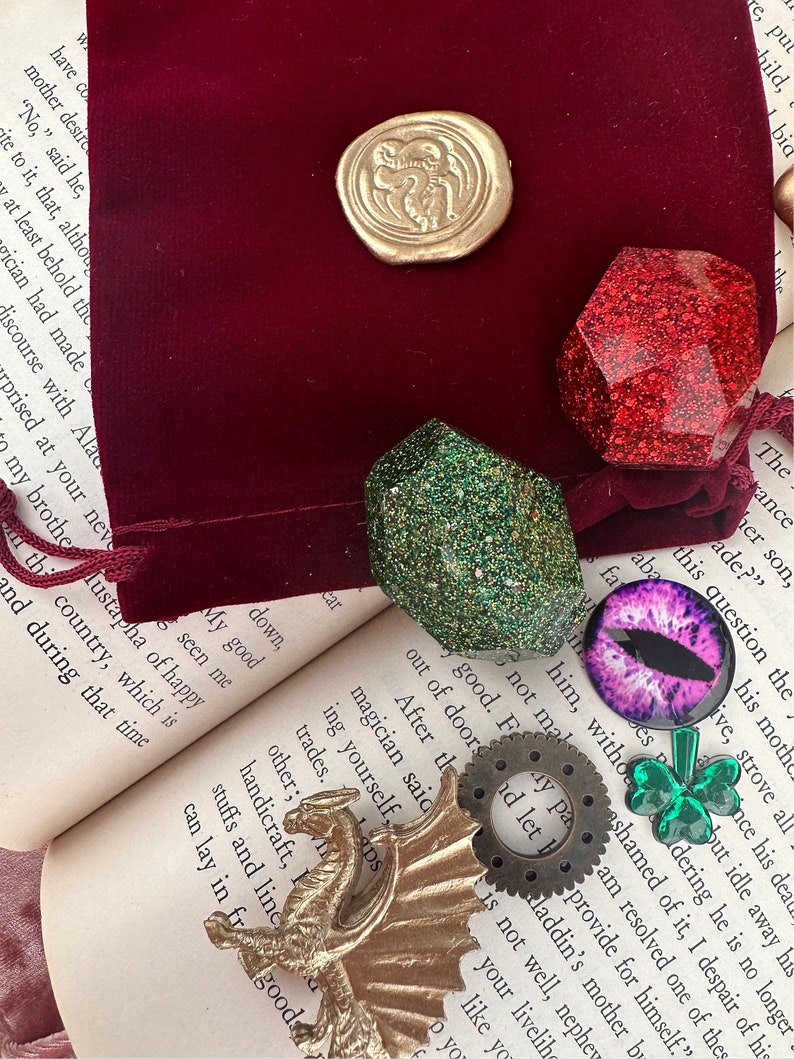 K&ouml;nnte beinhalten: Eine Sammlung von Fantasy-Accessoires, darunter ein goldener Drache, ein goldener Wachssiegel, ein roter Glitzer-Edelstein, ein gr&uuml;ner Glitzer-Edelstein, ein lila Augen-Cabochon, ein Zahnrad und ein gr&uuml;nes Kleeblatt. Diese Artikel sind auf einem offenen Buch und einem bordeauxroten Samtbeutel angeordnet.