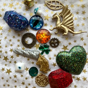 K&ouml;nnte beinhalten: Eine Sammlung dekorativer Verzierungen, darunter eine goldene Drachenfigur, ein gr&uuml;nes Glitzerherz und ein blauer Glitzerstein. Weitere Artikel sind Zahnr&auml;der, eine M&uuml;nze und ein vierbl&auml;ttriges Kleeblatt.