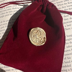 K&ouml;nnte beinhalten: Ein weinroter Samtbeutel mit Kordelzug und einem goldfarbenen Wachssiegel mit einem Drachendesign. Das Siegel ist auf der Oberfl&auml;che des Beutels befestigt. Der Beutel liegt auf einem offenen Buch.