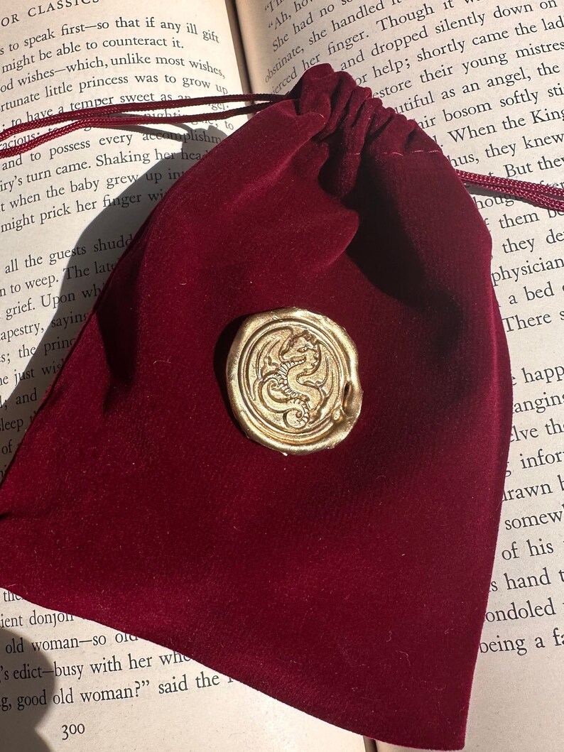 K&ouml;nnte beinhalten: Ein weinroter Samtbeutel mit Kordelzug und einem goldfarbenen Wachssiegel mit einem Drachendesign. Das Siegel ist auf der Oberfl&auml;che des Beutels befestigt. Der Beutel liegt auf einem offenen Buch.