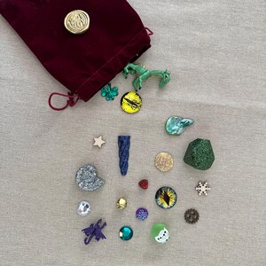K&ouml;nnte beinhalten: Ein bordeauxfarbener Samtbeutel mit Kordelzug und goldenem Siegel, umgeben von verschiedenen dekorativen Gegenst&auml;nden. Dazu geh&ouml;ren eine gr&uuml;ne Drachenfigur, ein gelb-schwarzer augenf&ouml;rmiger Anh&auml;nger und mehrere andere kleine, farbenfrohe Verzierungen.