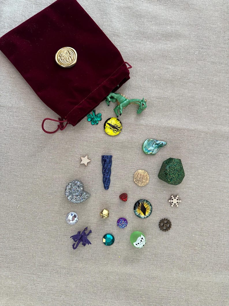 K&ouml;nnte beinhalten: Ein bordeauxfarbener Samtbeutel mit Kordelzug und goldenem Siegel, umgeben von verschiedenen dekorativen Gegenst&auml;nden. Dazu geh&ouml;ren eine gr&uuml;ne Drachenfigur, ein gelb-schwarzer augenf&ouml;rmiger Anh&auml;nger und mehrere andere kleine, farbenfrohe Verzierungen.