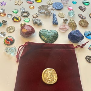 K&ouml;nnte beinhalten: Eine Sammlung von Fantasy-Verzierungen, darunter glitzernde Edelsteine, Miniaturdrachen und ein Samtbeutel mit einem goldenen Siegel. Das Sortiment umfasst verschiedene Formen und Farben, wie Herzen, Augen und Blumen, ideal f&uuml;r Bastelarbeiten oder Schmuckherstellung.