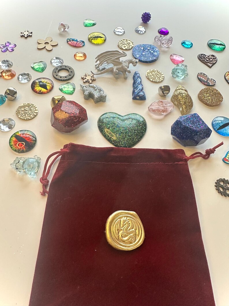 K&ouml;nnte beinhalten: Eine Sammlung von Fantasy-Verzierungen, darunter glitzernde Edelsteine, Miniaturdrachen und ein Samtbeutel mit einem goldenen Siegel. Das Sortiment umfasst verschiedene Formen und Farben, wie Herzen, Augen und Blumen, ideal f&uuml;r Bastelarbeiten oder Schmuckherstellung.