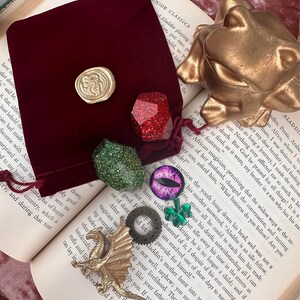 K&ouml;nnte beinhalten: Eine Sammlung von Fantasy-Accessoires, darunter eine goldene Drachenfigur, ein roter Samtbeutel mit goldenem Siegel und verschiedene edelsteinartige Anh&auml;nger in Rot, Gr&uuml;n und Lila. Eine goldene Froschstatue ist ebenfalls vorhanden.