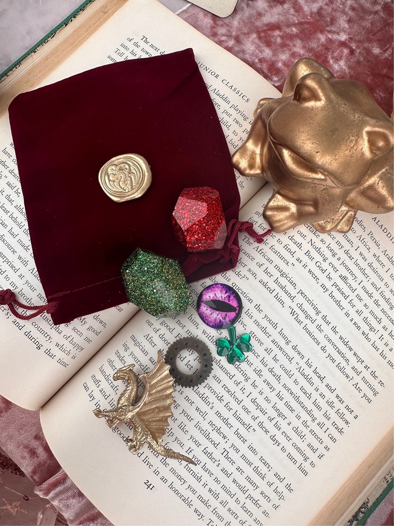 K&ouml;nnte beinhalten: Eine Sammlung von Fantasy-Accessoires, darunter eine goldene Drachenfigur, ein roter Samtbeutel mit goldenem Siegel und verschiedene edelsteinartige Anh&auml;nger in Rot, Gr&uuml;n und Lila. Eine goldene Froschstatue ist ebenfalls vorhanden.