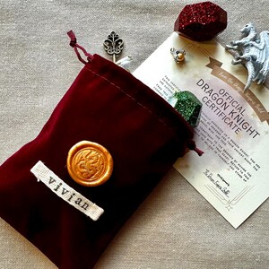 K&ouml;nnte beinhalten: Ein weinroter Samtbeutel mit goldenem Wachssiegel und dem Namen "vivian" auf einem wei&szlig;en Etikett. Ein Zertifikat lautet "Official Dragon Knight Certificate" mit einer Drachenfigur, einem roten Kristall und anderen kleinen Gegenst&auml;nden.