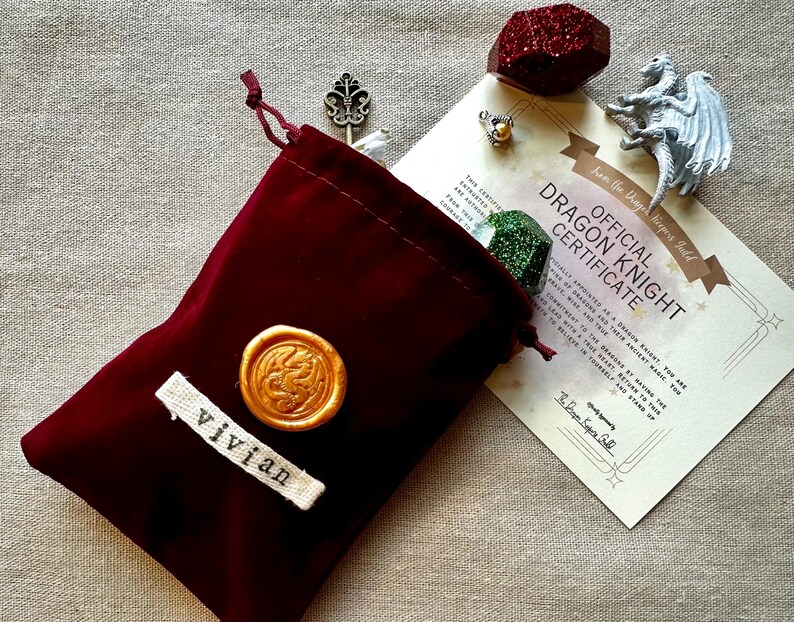 K&ouml;nnte beinhalten: Ein weinroter Samtbeutel mit goldenem Wachssiegel und dem Namen "vivian" auf einem wei&szlig;en Etikett. Ein Zertifikat lautet "Official Dragon Knight Certificate" mit einer Drachenfigur, einem roten Kristall und anderen kleinen Gegenst&auml;nden.