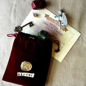K&ouml;nnte beinhalten: Ein burgunderfarbener Samtbeutel mit einem goldenen Siegel und dem Namen "oliver" auf einem wei&szlig;en Etikett. Das Bild zeigt auch ein Zertifikat, einen Schl&uuml;ssel, eine Drachenfigur und edelsteinartige Objekte in Rot und Gr&uuml;n.