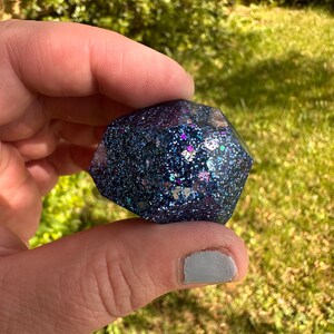 K&ouml;nnte beinhalten: Ein dunkelblaues, facettenreiches Objekt aus Kunstharz mit eingebetteten Glitzerpartikeln in Blau-, Lila- und Silbert&ouml;nen. Das Objekt wird zwischen Fingern vor einem verschwommenen gr&uuml;nen Hintergrund gehalten.