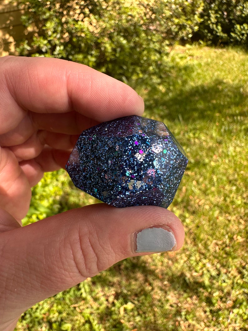 K&ouml;nnte beinhalten: Ein dunkelblaues, facettenreiches Objekt aus Kunstharz mit eingebetteten Glitzerpartikeln in Blau-, Lila- und Silbert&ouml;nen. Das Objekt wird zwischen Fingern vor einem verschwommenen gr&uuml;nen Hintergrund gehalten.